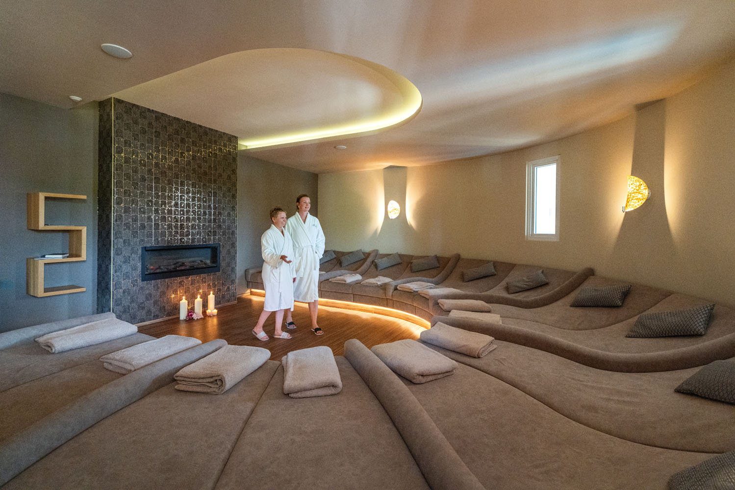 Zwei Personen in weißen Bademänteln stehen in einem modernen Spa-Ruheraum mit geschwungenen Liegestühlen, gerollten Handtüchern, einem Kamin und sanfter Beleuchtung. Die Atmosphäre wirkt ruhig und einladend.