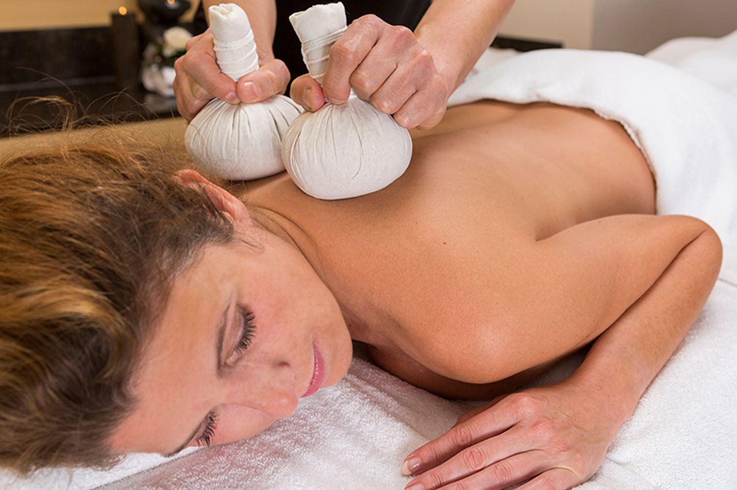 Eine Frau liegt mit dem Gesicht nach unten auf einem Massagetisch, bedeckt mit einem weißen Handtuch. Eine Person massiert mit zwei Kräuterkompressenkugeln ihren oberen Rücken und ihre Schultern. Die Umgebung wirkt ruhig und entspannend.