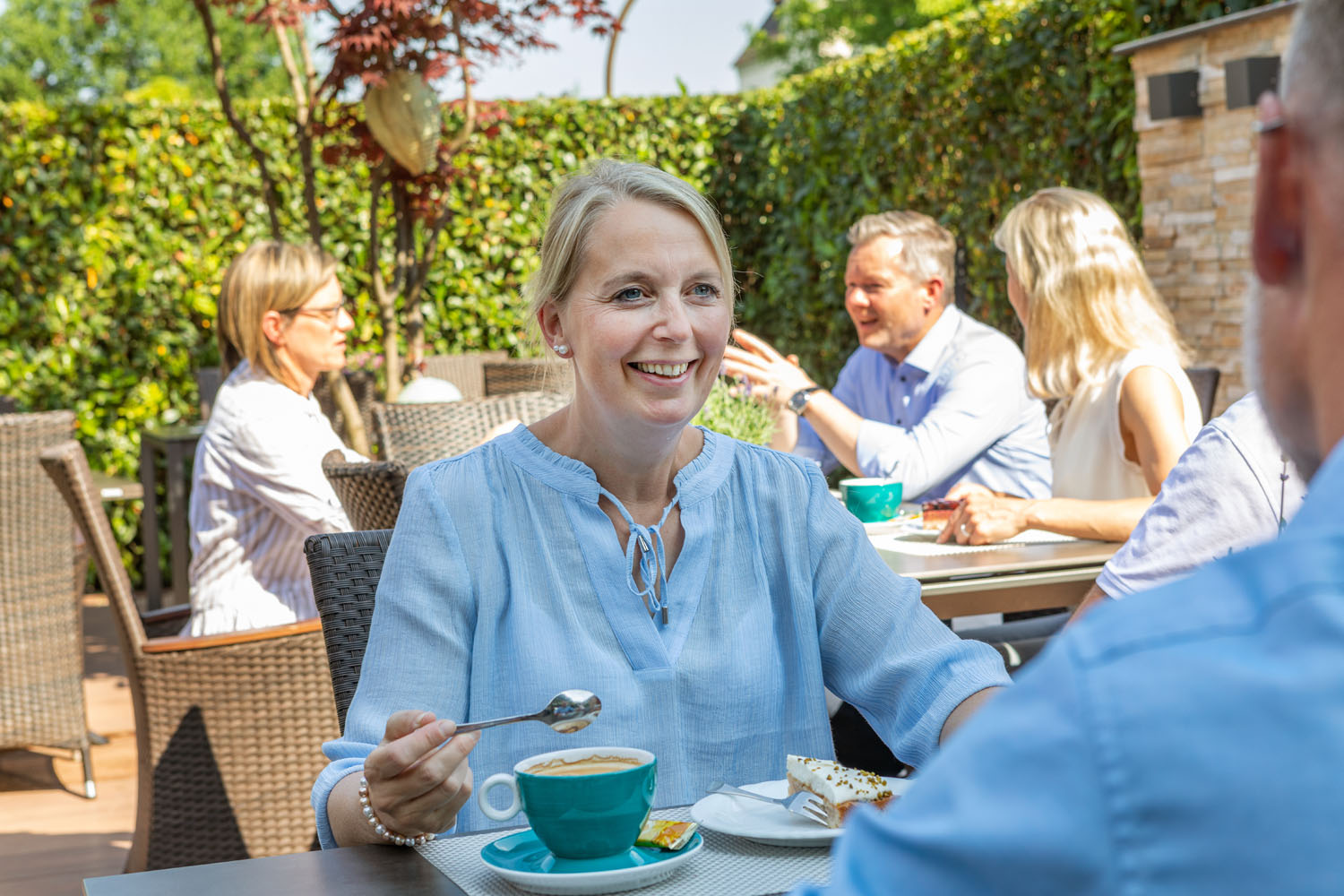 Eine Frau lächelt, während sie einen Löffel hält und mit einer Tasse Kaffee und einem Dessert an einem Cafétisch im Freien sitzt. Im Hintergrund unterhalten sich andere Menschen und genießen ihre Getränke an einem sonnigen Tag.