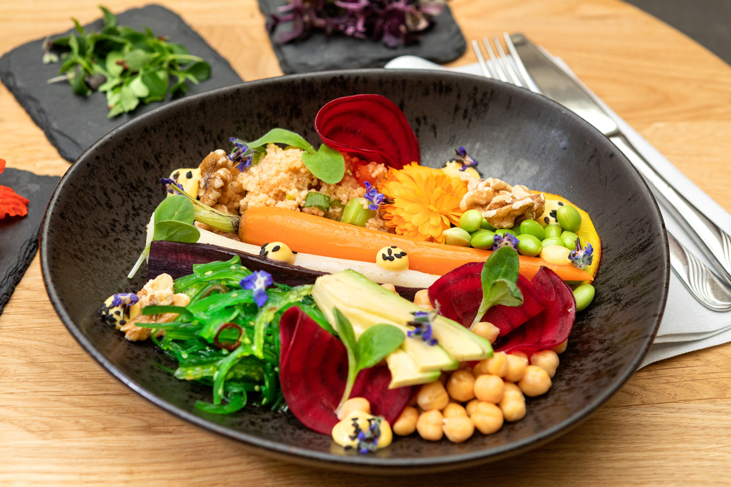 Ein farbenfroher Salat in einer schwarzen Schale mit Rübenscheiben, Avocado, Kichererbsen, Karotten, Edamame, Algen, Mikrogrün, Walnüssen, Quinoa und essbaren Blüten wird kunstvoll auf einem Holztisch mit Besteck daneben angerichtet.