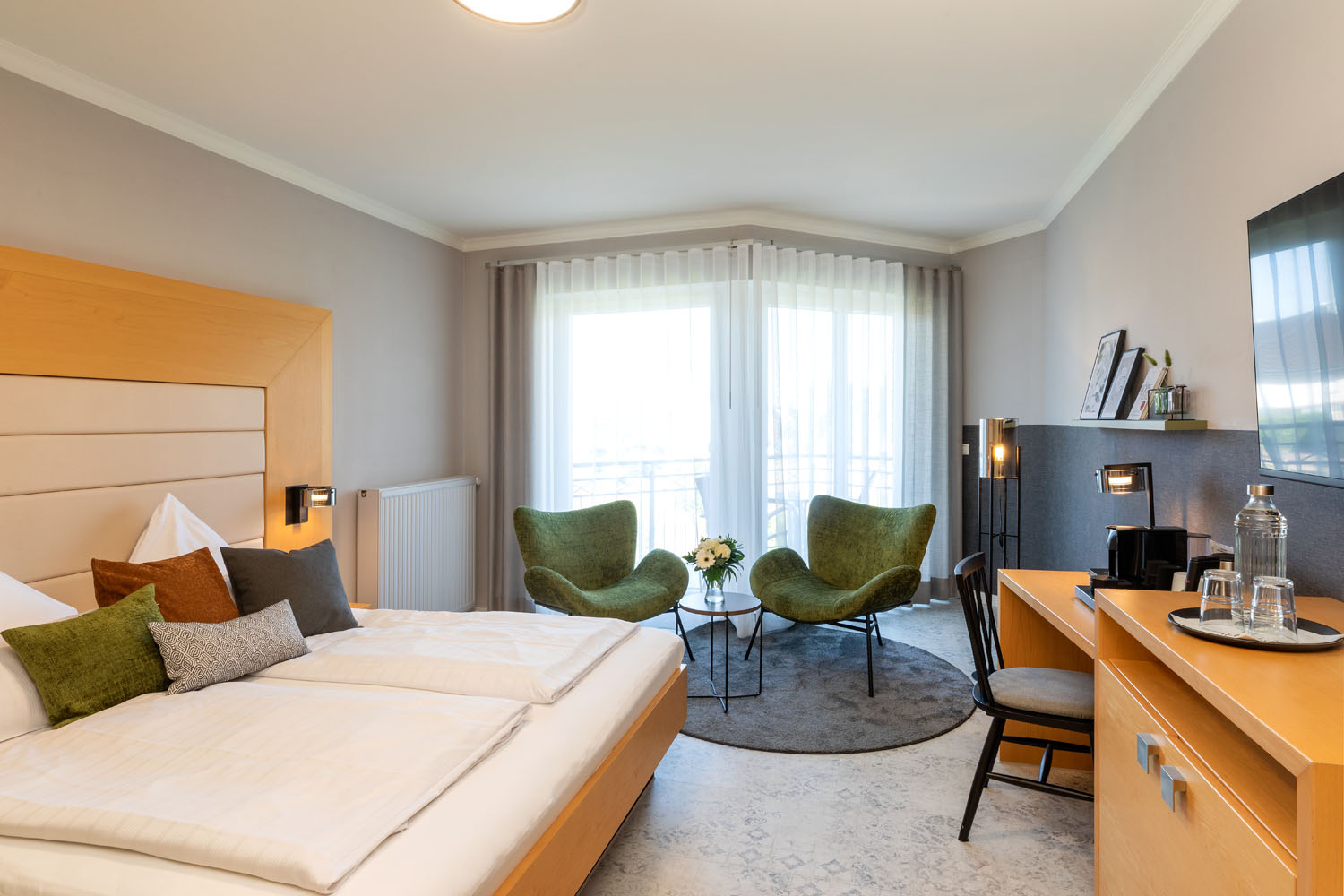 Ein modernes Hotelzimmer mit einem großen Bett, grünen Akzentstühlen um einen kleinen Tisch, einem Schreibtisch mit Fernseher und einem Fenster mit durchsichtigen Vorhängen, die natürliches Licht hereinlassen.