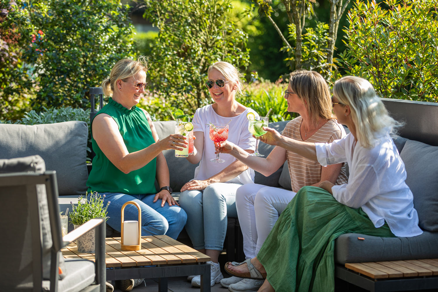 Vier Frauen sitzen auf einer Terrasse im Freien, lächeln und stoßen mit bunten Getränken an. Sie sind von Grün und Sonnenlicht umgeben, was eine fröhliche und entspannte Atmosphäre schafft.