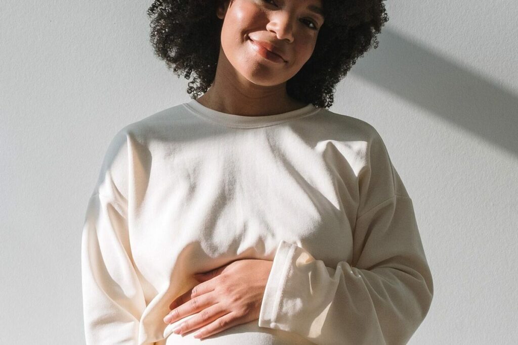 Eine lächelnde Frau mit lockigem Haar steht in einem hellen, langärmeligen Hemd vor einer weißen Wand und stützt ihre Hand sanft auf ihren Bauch. Das Sonnenlicht wirft weiche Schatten hinter sie.
