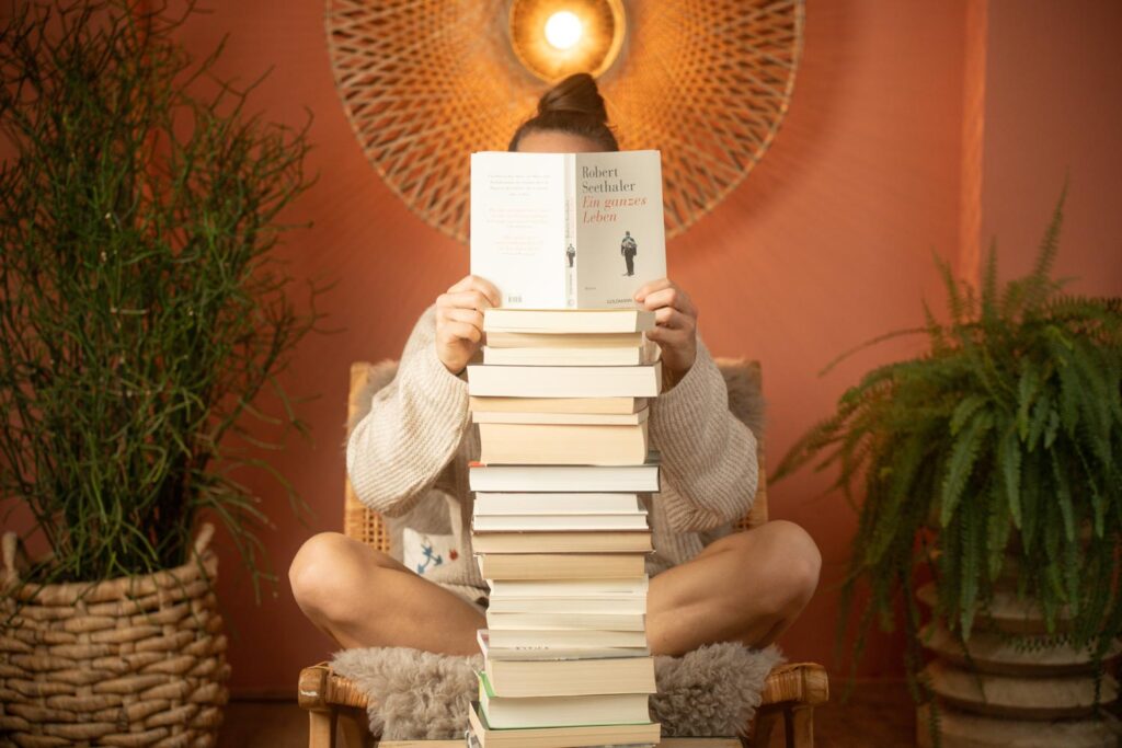 Eine Person sitzt im Schneidersitz auf einem gemütlichen Stuhl, hält ein aufgeschlagenes Buch in der Hand und verbirgt ihr Gesicht dahinter. Vor ihr steht ein hoher Stapel Bücher. Im Hintergrund sind Pflanzen und eine warme, dekorative Wandlampe zu sehen.