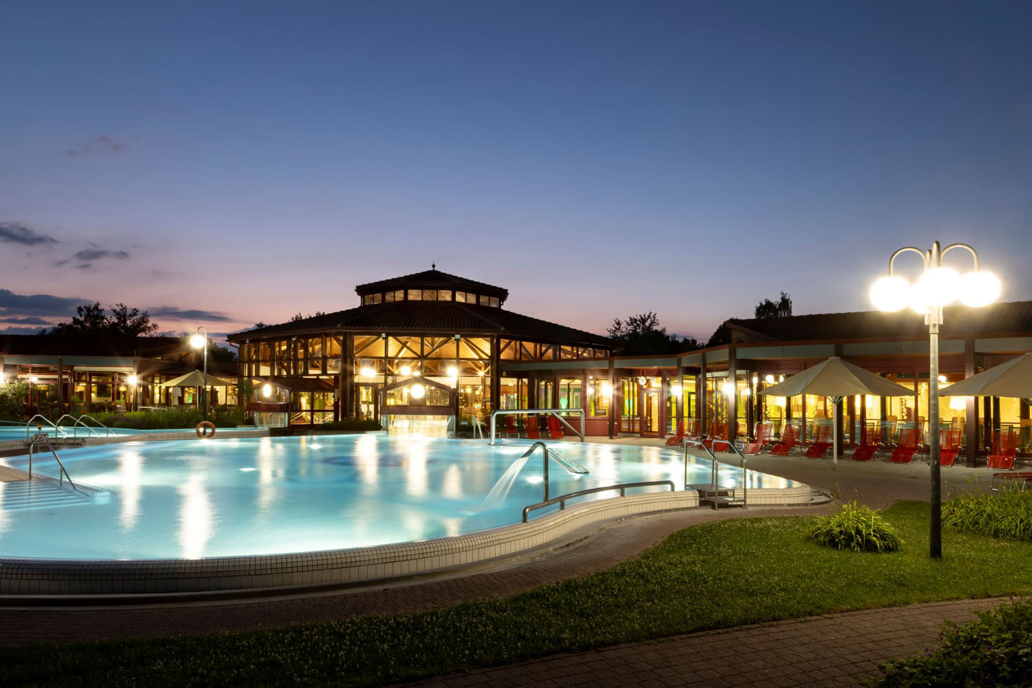 Ein großer Swimmingpool im Freien mit leuchtenden Lichtern, Liegestühlen und Sonnenschirmen vor einem modernen Gebäude in der Abenddämmerung, umgeben von Grünflächen.