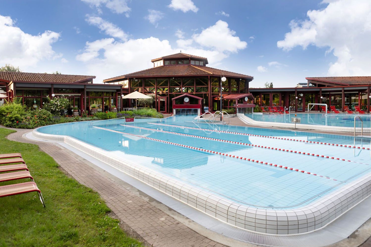 Außenschwimmbad mit Bahntrennung, umgeben von Liegestühlen und Grünanlagen, vor einem großen Holzgebäude mit Glasfenstern unter einem teilweise bewölkten blauen Himmel.