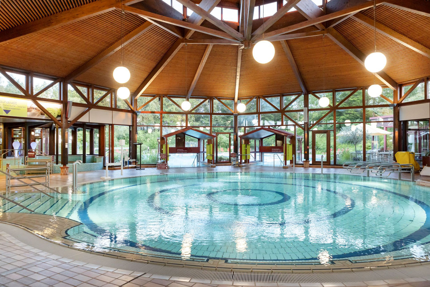 Rundes Hallenbad mit klarem Wasser, umgeben von Fliesenböden und Holzbalken. Große Fenster lassen natürliches Licht herein und geben den Blick auf die grüne Umgebung frei. Darüber hängen helle Deckenleuchten.