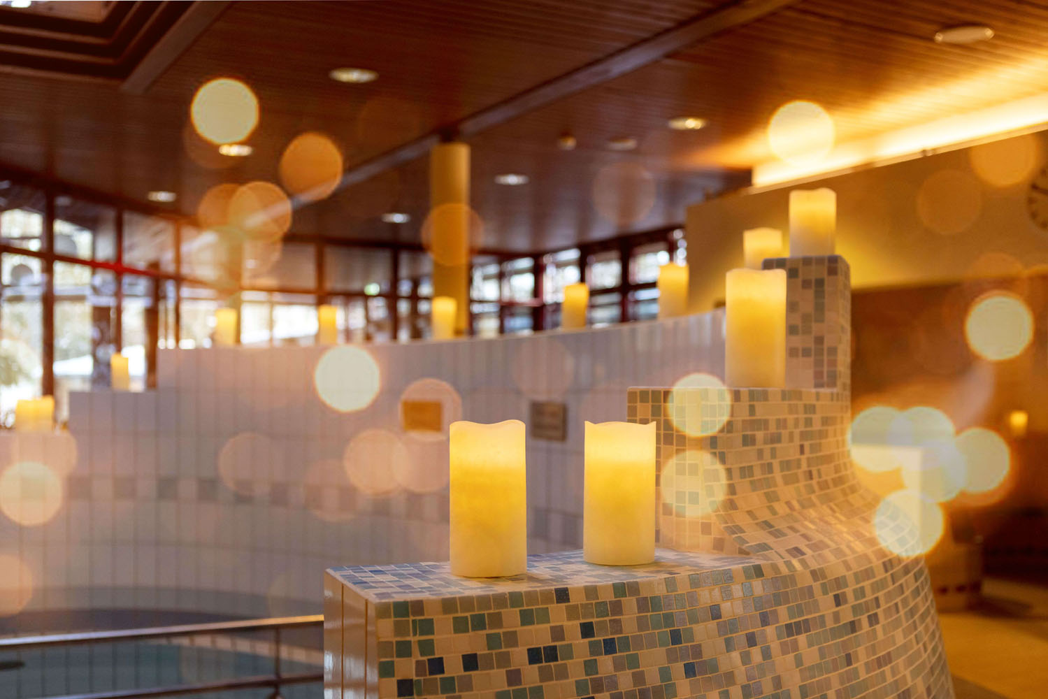 Eine Reihe von brennenden Stumpenkerzen steht auf einem Kachelsims in einem sanft beleuchteten Raum, in dem warme Bokeh-Lichteffekte eine gemütliche, ruhige Atmosphäre schaffen. Im Hintergrund sind große Fenster und eine Holzdecke zu sehen.