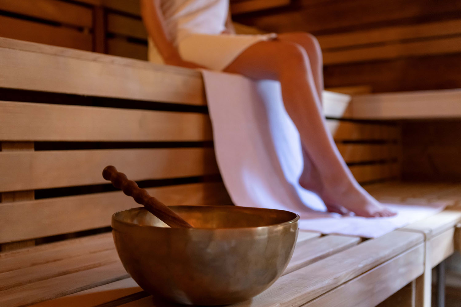 Eine in ein weißes Handtuch gehüllte Person sitzt auf einer Holzbank in einer Sauna, unter der ein Handtuch liegt. Im Vordergrund ruht eine Klangschale auf dr Saunabank.