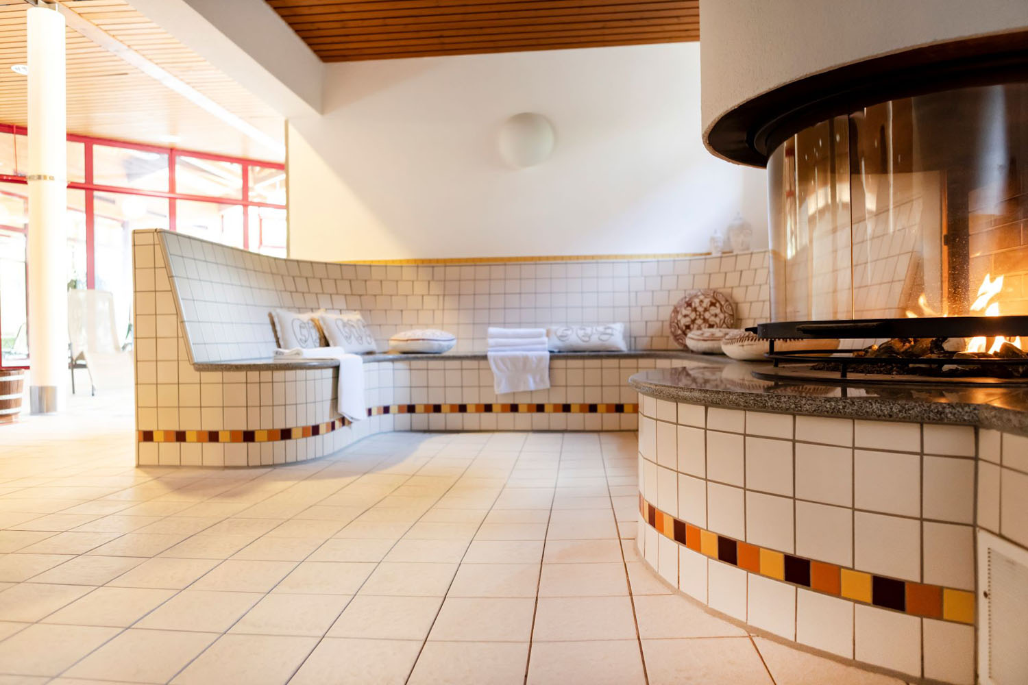 Ein moderner Saunaraum mit gefliesten Bänken, gefalteten Handtüchern und einem runden Glaskamin. Der Raum ist hell, sauber und einladend, mit warmer Beleuchtung und dekorativen Kachelakzenten.