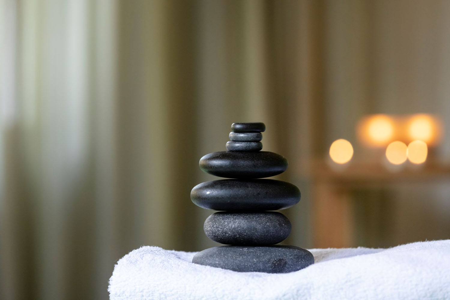 Ein Stapel glatter schwarzer Steine auf einem weißen Handtuch, mit einem verschwommenen Hintergrund mit warmen, leuchtenden Lichtern, die eine ruhige und entspannende Atmosphäre schaffen.