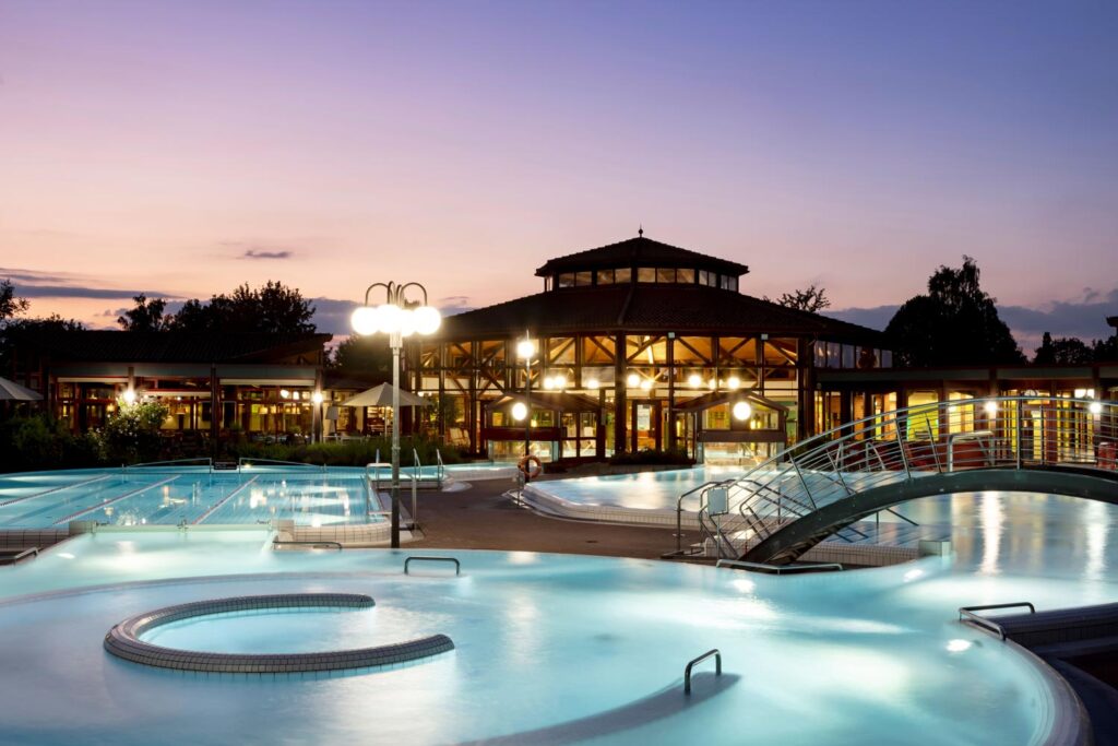 Vor dem großen, runden Gebäude der Sonnenhof Therme sind in der Abenddämmerung Außenbecken mit leuchtenden Lichtern und eine kleine Brücke zu sehen. Der Himmel ist rosa und lila, und Bäume umrahmen die entspannende, beleuchtete Szene.