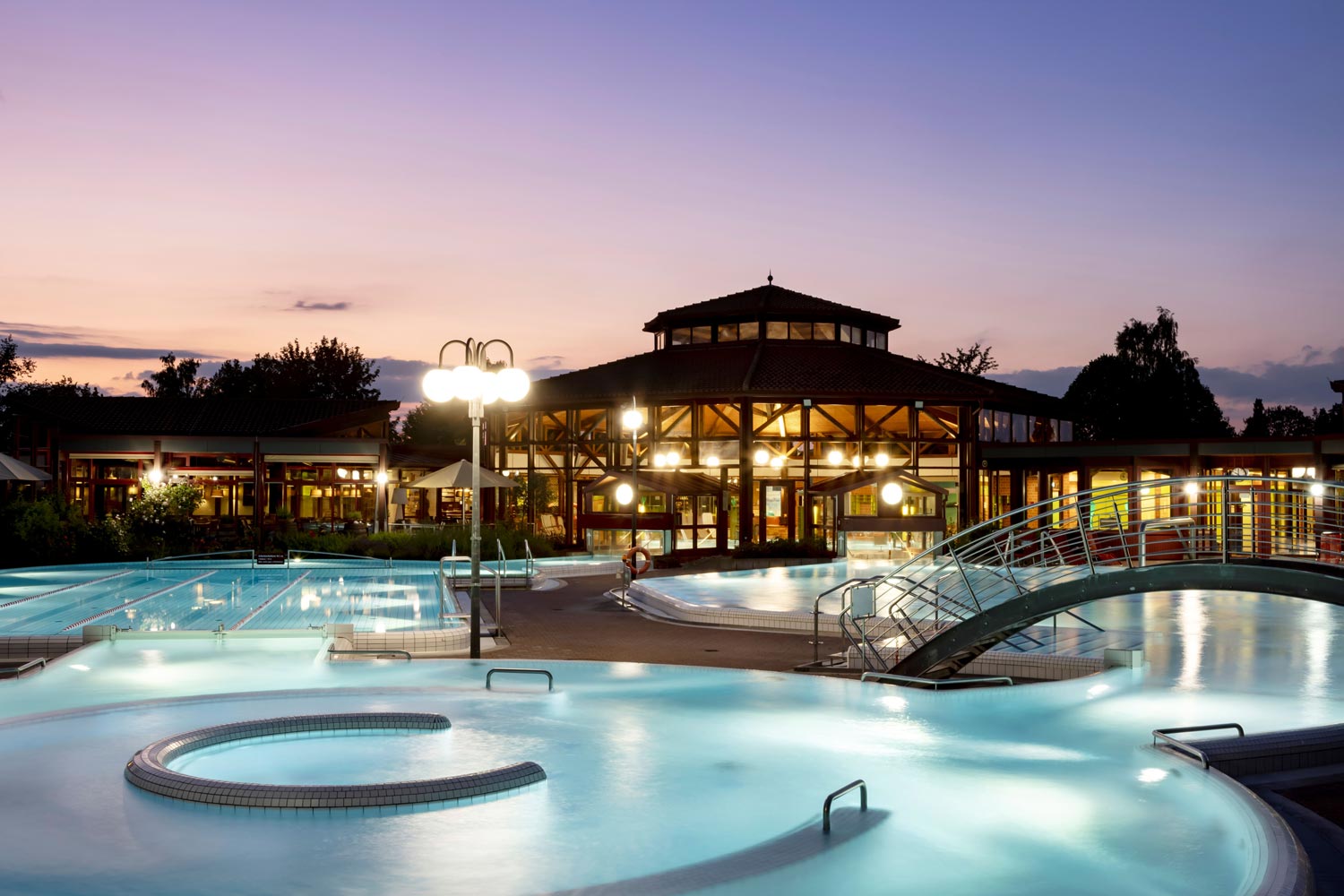 Vor dem großen, runden Gebäude der Sonnenhof Therme sind in der Abenddämmerung Außenbecken mit leuchtenden Lichtern und eine kleine Brücke zu sehen. Der Himmel ist rosa und lila, und Bäume umrahmen die entspannende, beleuchtete Szene.