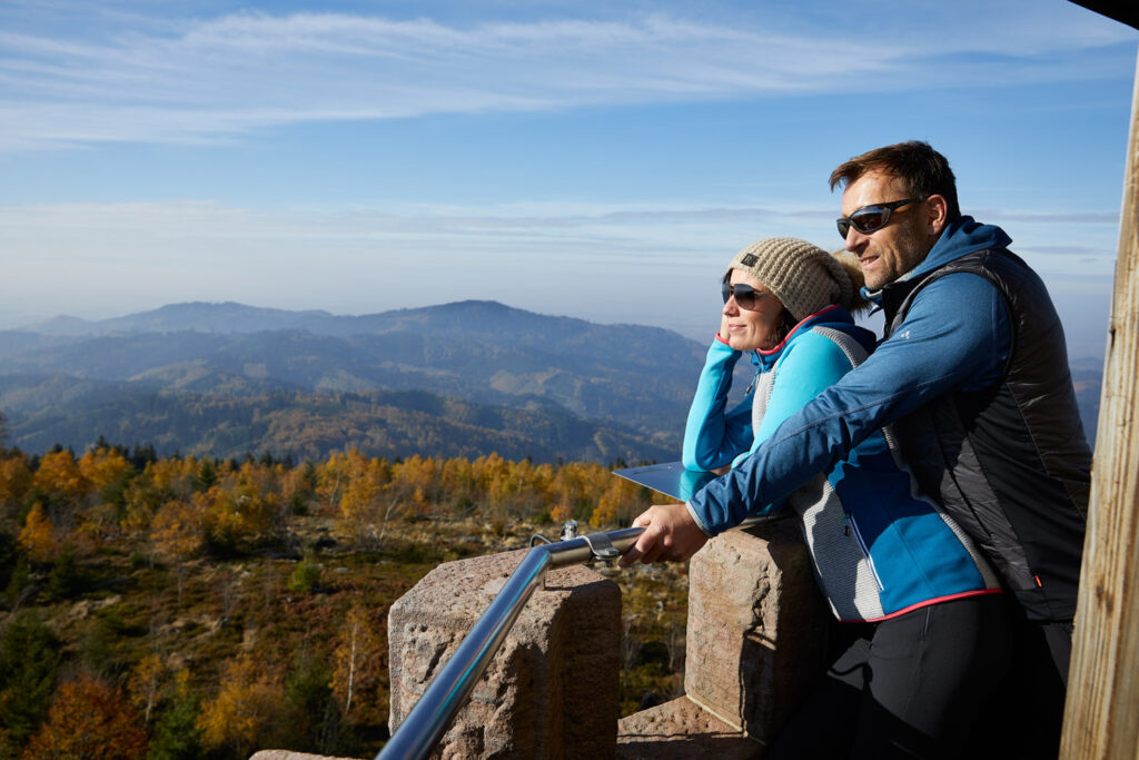 Ein Paar mit Sonnenbrille und warmer Kleidung steht auf einem steinernen Balkon und genießt die Aussicht auf die Berge mit ihren sanften Hügeln und herbstlichen Bäumen unter blauem Himmel in der Ortenau.