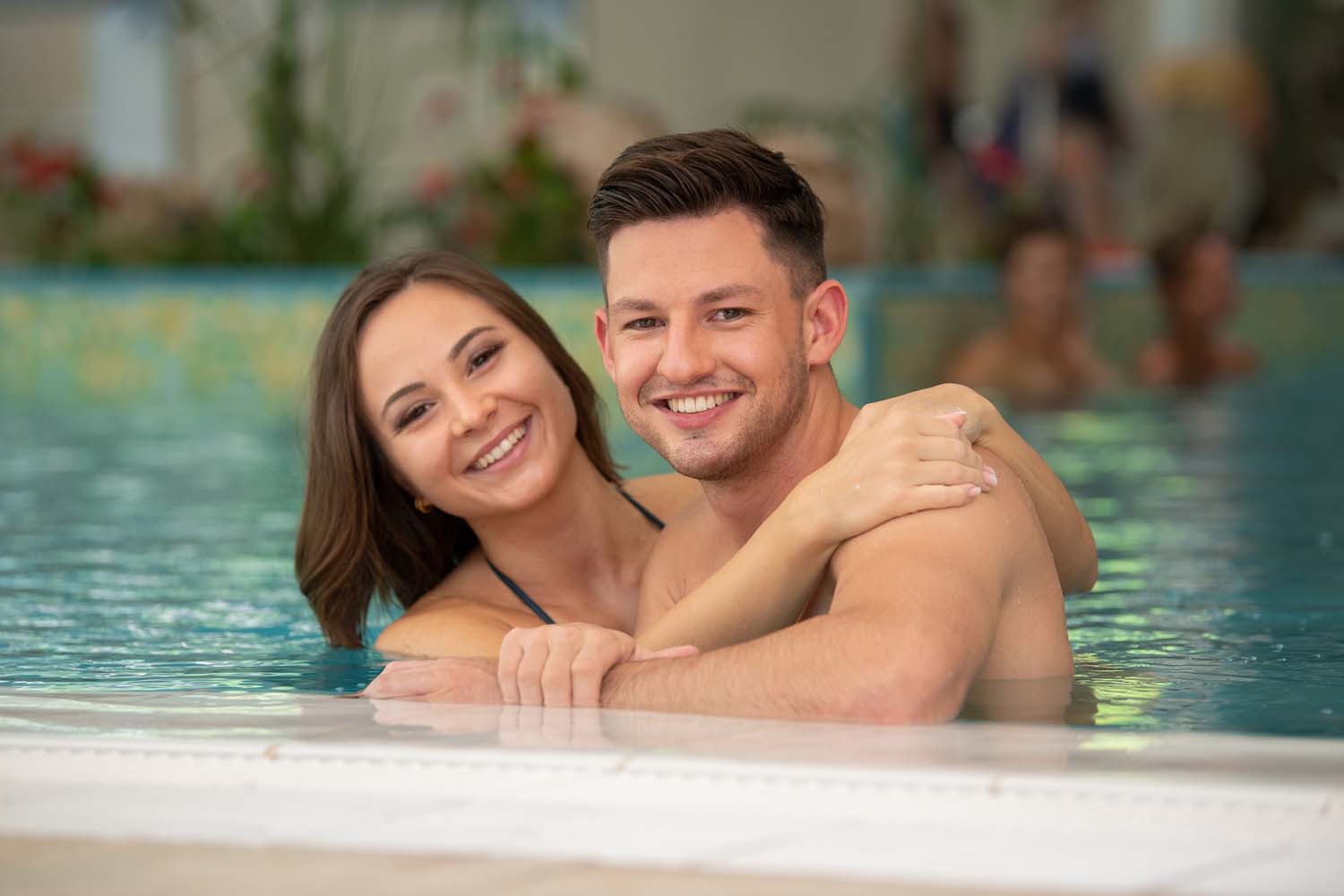 Ein lächelndes junges Paar umarmt sich in einem Schwimmbad und ruht sich am Beckenrand aus. Die Frau hat ihre Arme um den Mann gelegt, und beide schauen in die Kamera. Der Pool hat einen unscharfen Hintergrund mit Pflanzen und anderen Menschen.