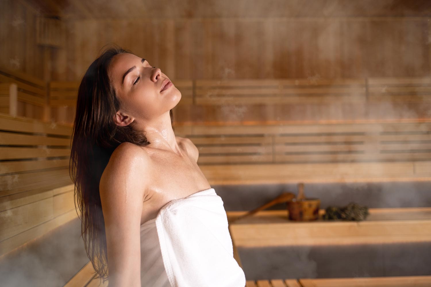 Eine in ein Handtuch gehüllte Frau entspannt sich mit geschlossenen Augen in einer Holzsauna, umgeben von Dampf und warmem Licht.