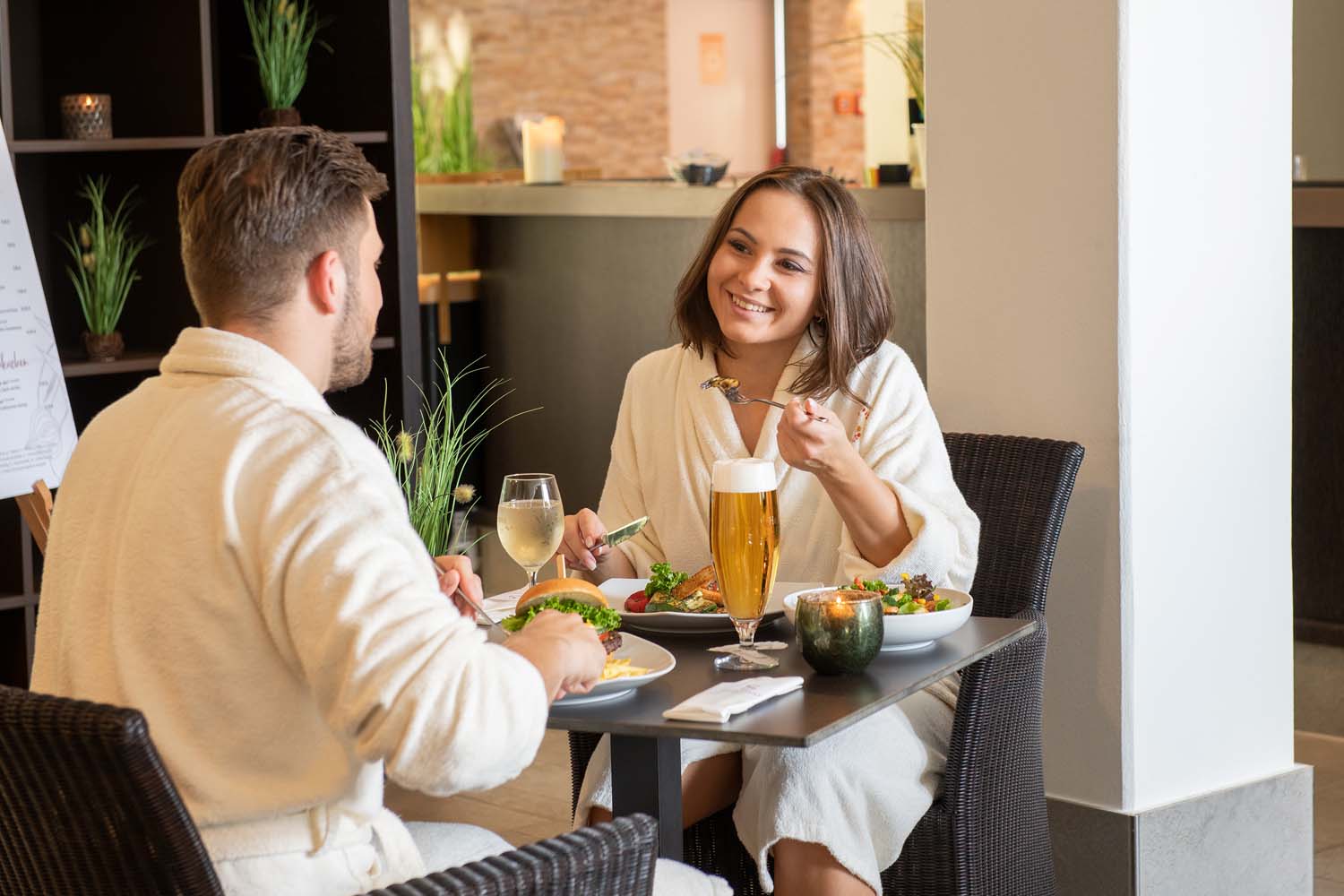 Ein Mann und eine Frau in weißen Gewändern sitzen an einem Tisch in einem Restaurant und genießen eine Mahlzeit mit Getränken, darunter Bier und Wein. Die Frau lächelt, während sie eine Gabel hält, und beide haben Teller mit Essen vor sich.