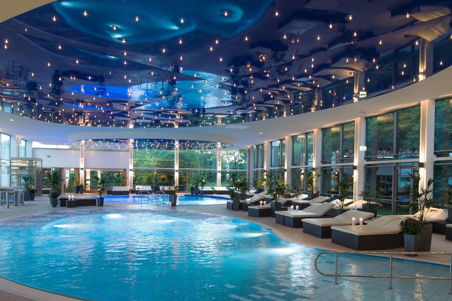 Luxuriöser Innenpool mit Liegestühlen, großen Fenstern mit Blick ins Grüne und einer auffallend blauen, sternförmig beleuchteten Decke, die sich im Wasser spiegelt. Der Raum ist modern, geräumig und einladend.