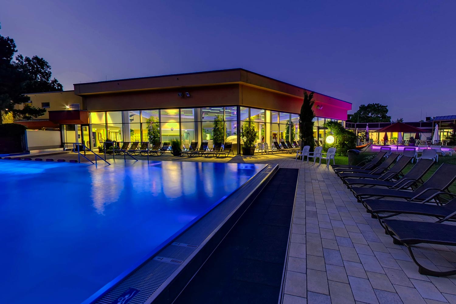 Mit blauen Lichtern beleuchtetes Freibad in der Abenddämmerung, umgeben von Liegestühlen, mit einem modernen gläsernen Gebäude, das im Hintergrund mit gelber Innenbeleuchtung leuchtet. Bäume und weitere Sitzgelegenheiten sind unter dem Dämmerungshimmel zu sehen.
