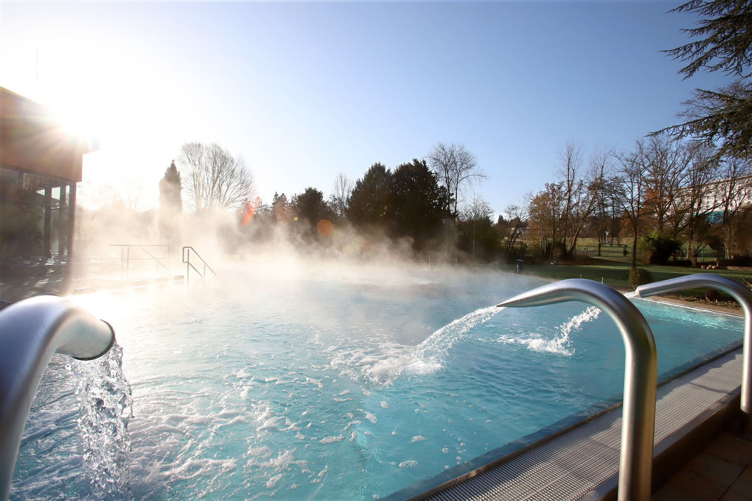 Ein Außenschwimmbad mit Wasserdüsen, die im morgendlichen Sonnenlicht Dampf ausstoßen. Bäume und eine Rasenfläche umgeben den Pool, der Himmel ist klar und blau.