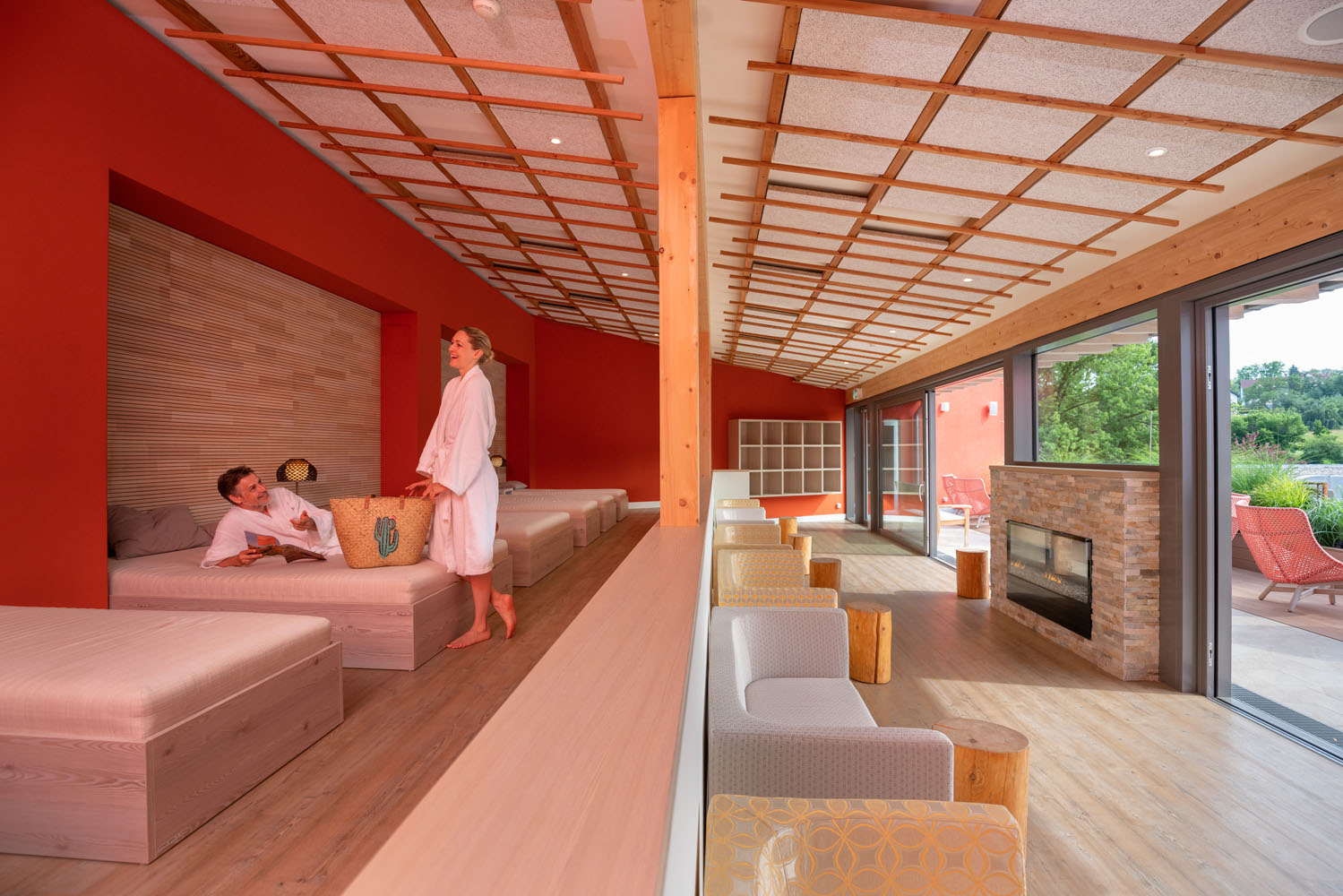Zwei Personen in weißen Bademänteln entspannen sich in einer modernen Spa-Lounge mit roten Wänden, großen Betten und Holzakzenten. Der Raum verfügt über einen gemütlichen Kamin, Sessel und raumhohe Fenster mit Blick ins Grüne.