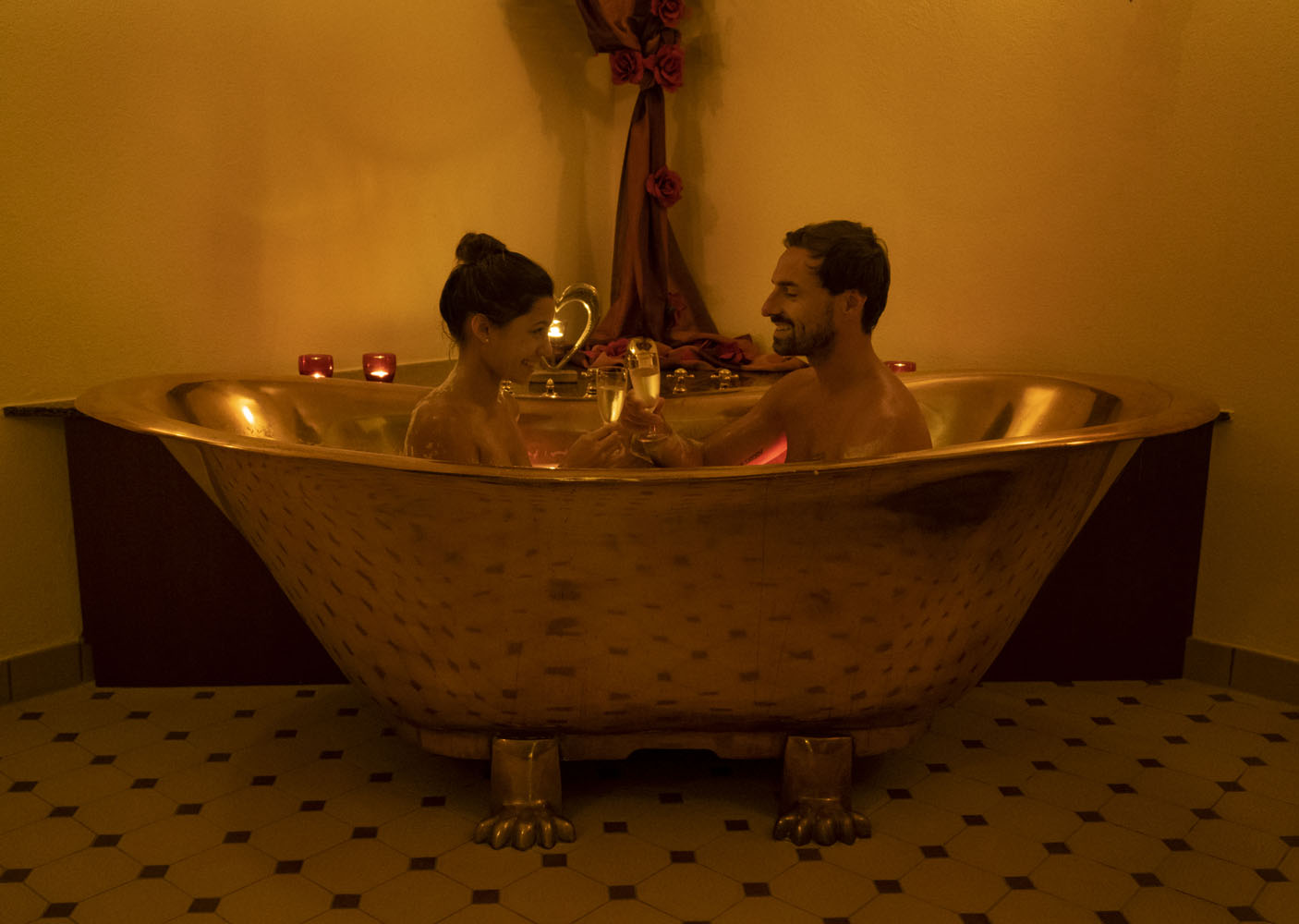 Ein Paar entspannt sich in einer goldenen Klauenfuß-Badewanne, umgeben von Kerzenlicht. Sie stehen sich gegenüber und stoßen mit Getränken an, wodurch eine romantische Atmosphäre in einem warm beleuchteten Raum entsteht.