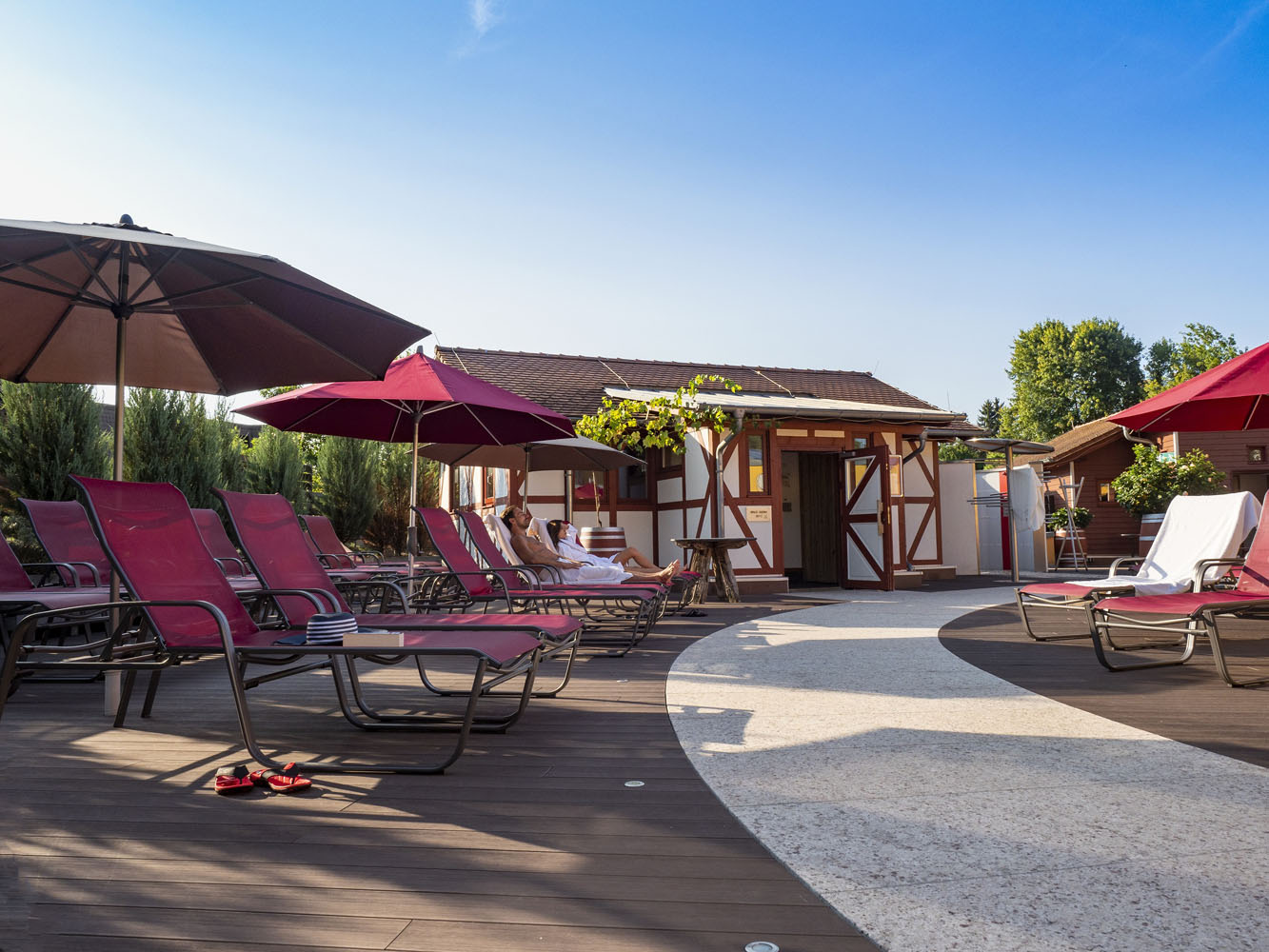 Wellnessbereich im Freien mit roten Liegestühlen, großen Sonnenschirmen und einem Holzsteg. Im Hintergrund sind ein Fachwerkhaus und grüne Bäume unter einem klaren blauen Himmel zu sehen.