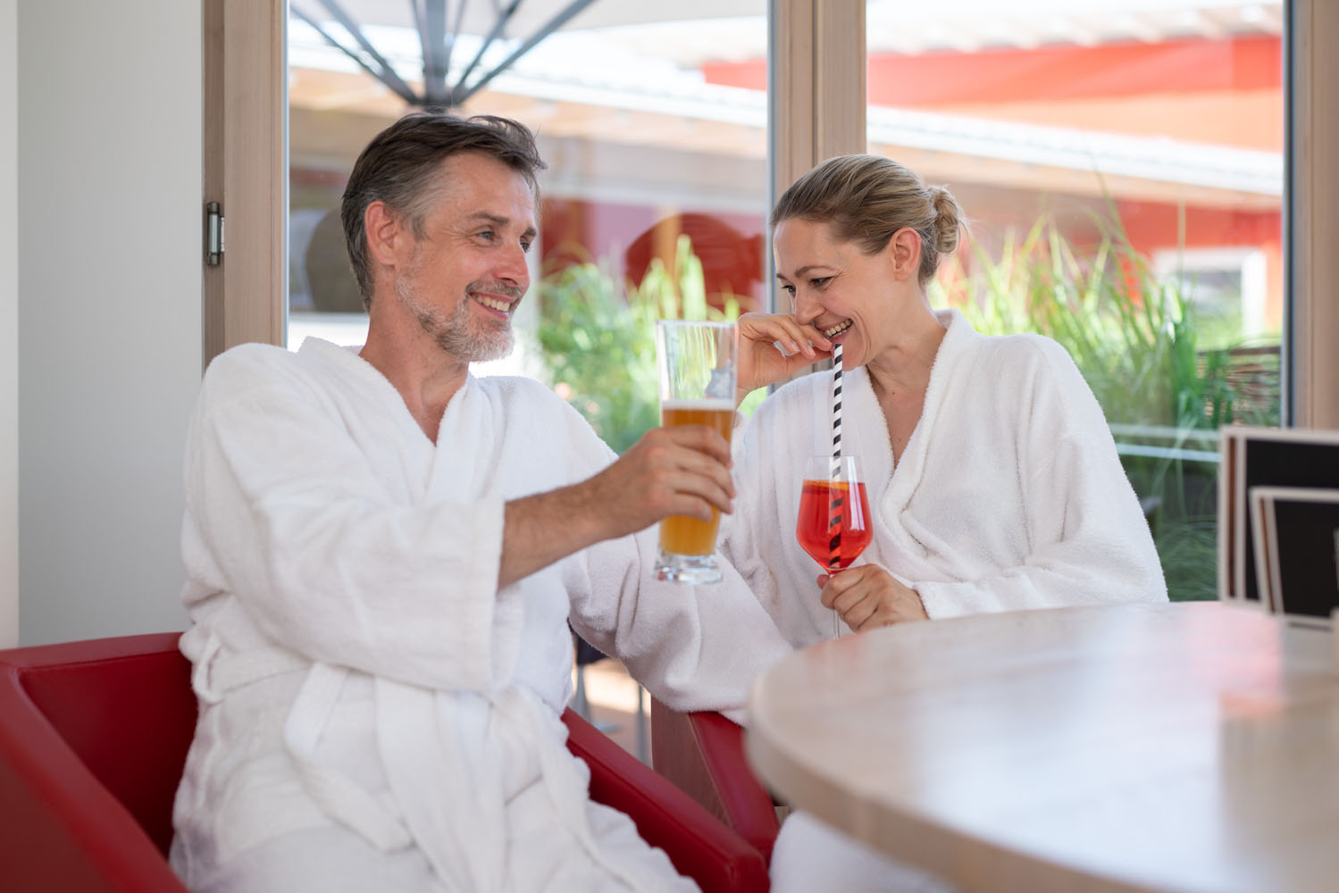 Ein Mann und eine Frau in weißen Bademänteln sitzen in einem hellen, modernen Raum mit großen Fenstern lächelnd an einem runden Tisch und genießen einen Drink; er hält ein Bier, sie einen rosa Cocktail mit Strohhalm.