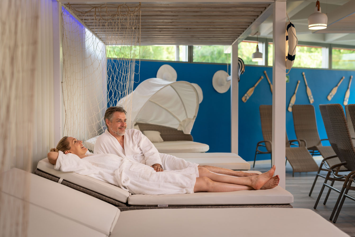 Ein lächelnder Mann und eine lächelnde Frau in weißen Bademänteln entspannen sich auf Liegestühlen in einem hellen, maritim gestalteten Wellnessbereich mit blauen Wänden, Paddeln und Korbliegen im Hintergrund.