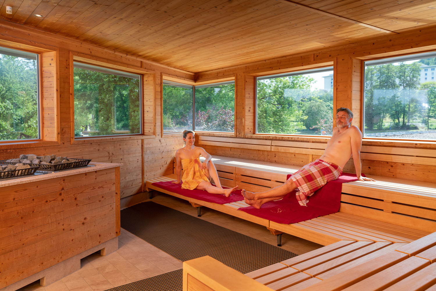 Zwei Personen entspannen sich auf Holzbänken in einem geräumigen Saunaraum mit großen Fenstern, die natürliches Licht und eine schöne Aussicht auf die Umgebung bieten. Beide sind in Handtücher gehüllt und wirken ruhig und bequem.