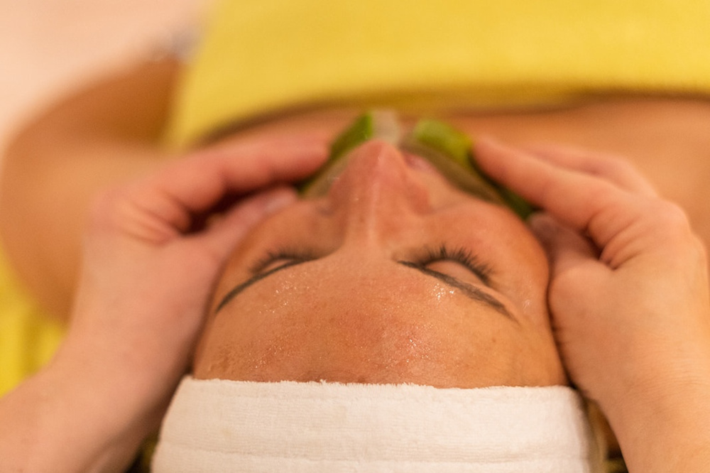 Eine Person mit geschlossenen Augen erhält eine entspannende Gesichtsmassage, indem sie sich mit einem weißen Stirnband und einem gelben Handtuch hinlegt, während zwei Hände sanft auf ihre Schläfen drücken.