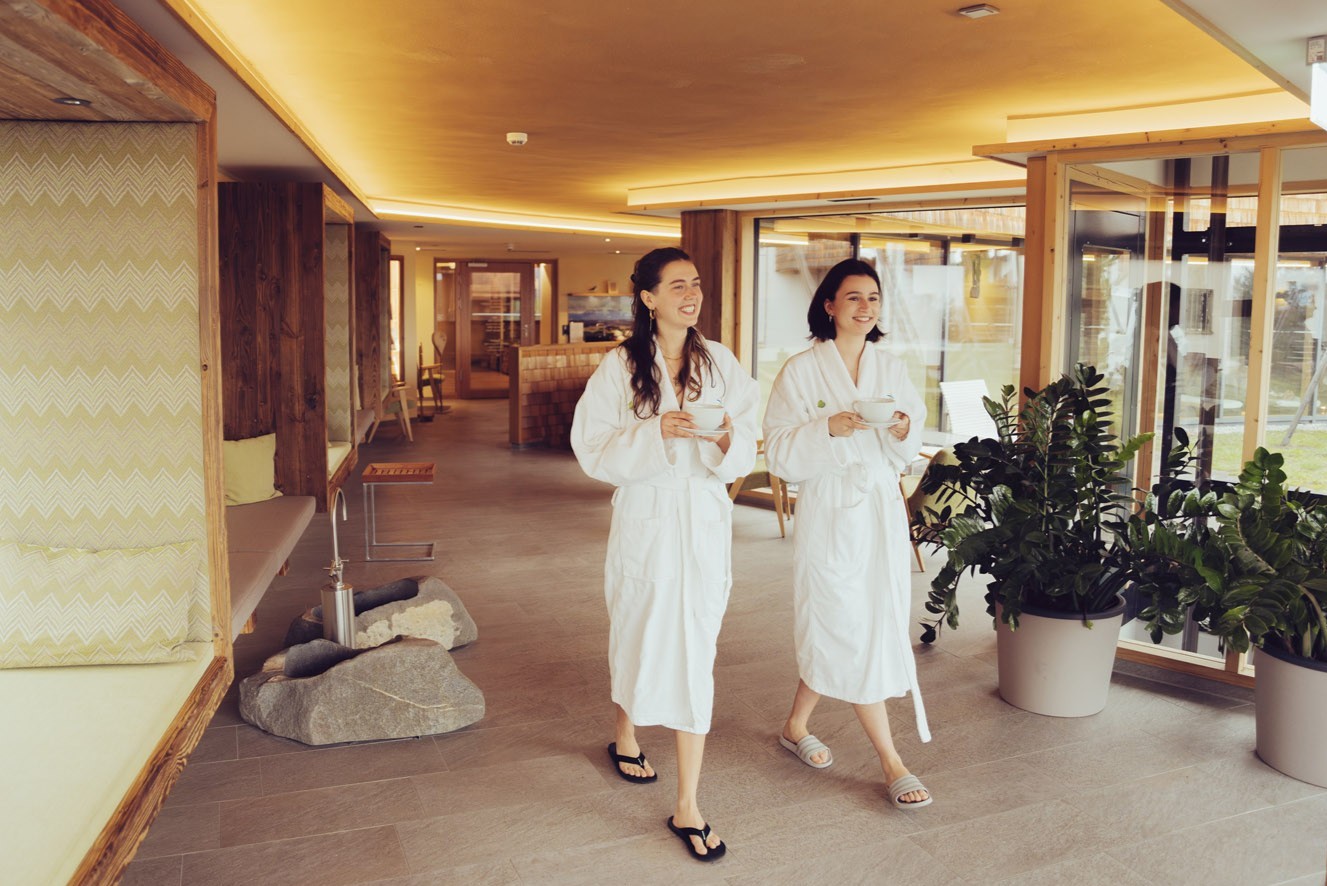 Zwei Frauen in weißen Bademänteln und Sandalen gehen lächelnd und mit Tassen in der Hand durch eine moderne, gut beleuchtete Spa-Lounge. Die Lounge ist mit Pflanzen, Holzakzenten und bequemen Sitzgelegenheiten ausgestattet.
