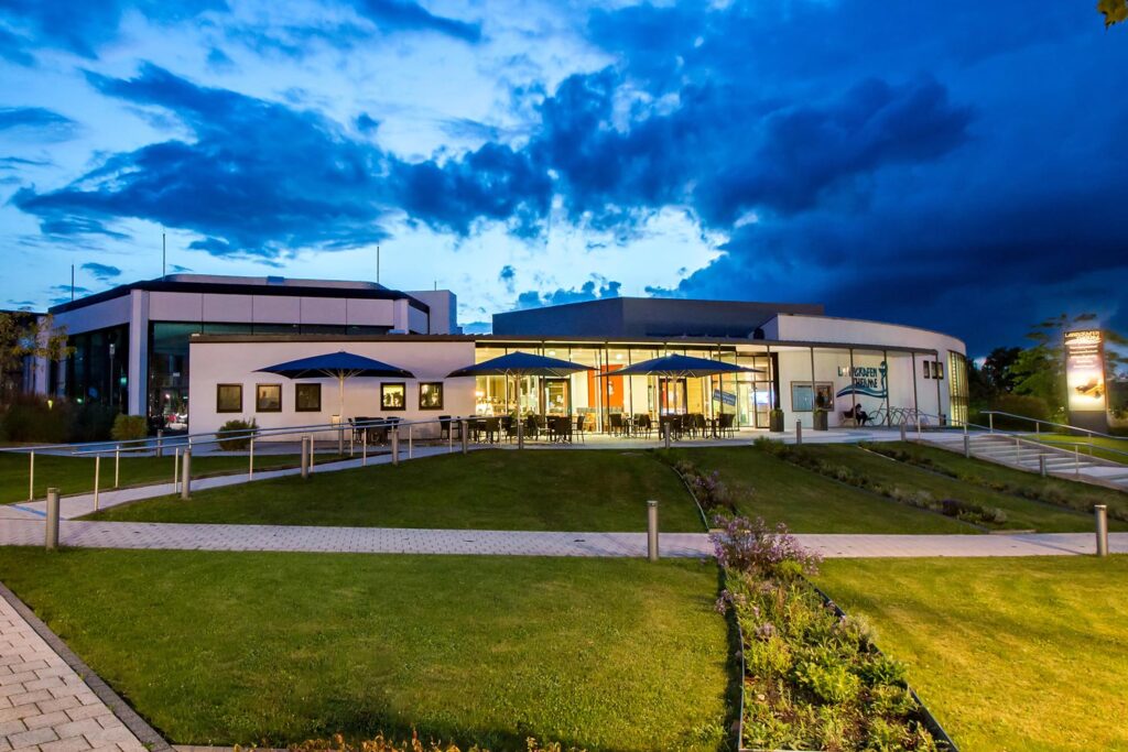 Modernes Gebäude mit großen Fenstern und Sitzplätzen im Freien unter blauen Sonnenschirmen, umgeben von Rasenflächen und Wegen, fotografiert in der Abenddämmerung mit dramatischen Wolken und lebendiger Beleuchtung.