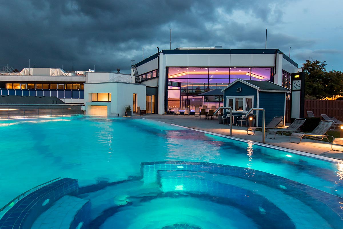 Ein modernes Gebäude mit großen Fenstern wird in der Abenddämmerung mit violetten Lichtern beleuchtet, daneben befinden sich ein leuchtender Außenpool und ein Whirlpool, über dem dunkle, dramatische Wolken hängen, und am Pool stehen Liegestühle.