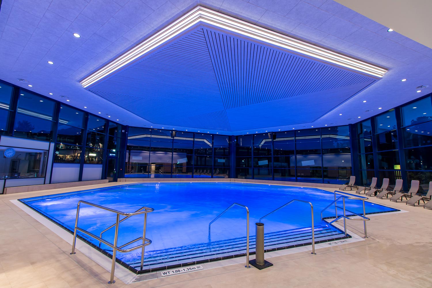 Großes Hallenbad mit blauer Beleuchtung, moderner geometrischer Decke, Handläufen aus Edelstahl und Reihen von Liegestühlen auf der rechten Seite. Vom Boden bis zur Decke reichende Fenster umgeben den Poolbereich.