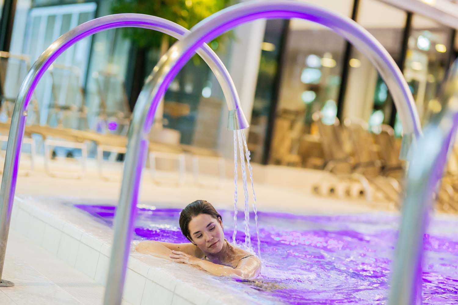 Eine Frau entspannt sich in einem Hallenbad mit violetter Beleuchtung. Sie stützt ihre Arme auf den Beckenrand, während Wasser aus einem gebogenen Metallbrunnen auf ihre Schultern fließt. Im Hintergrund sind Liegestühle und große Fenster zu sehen.