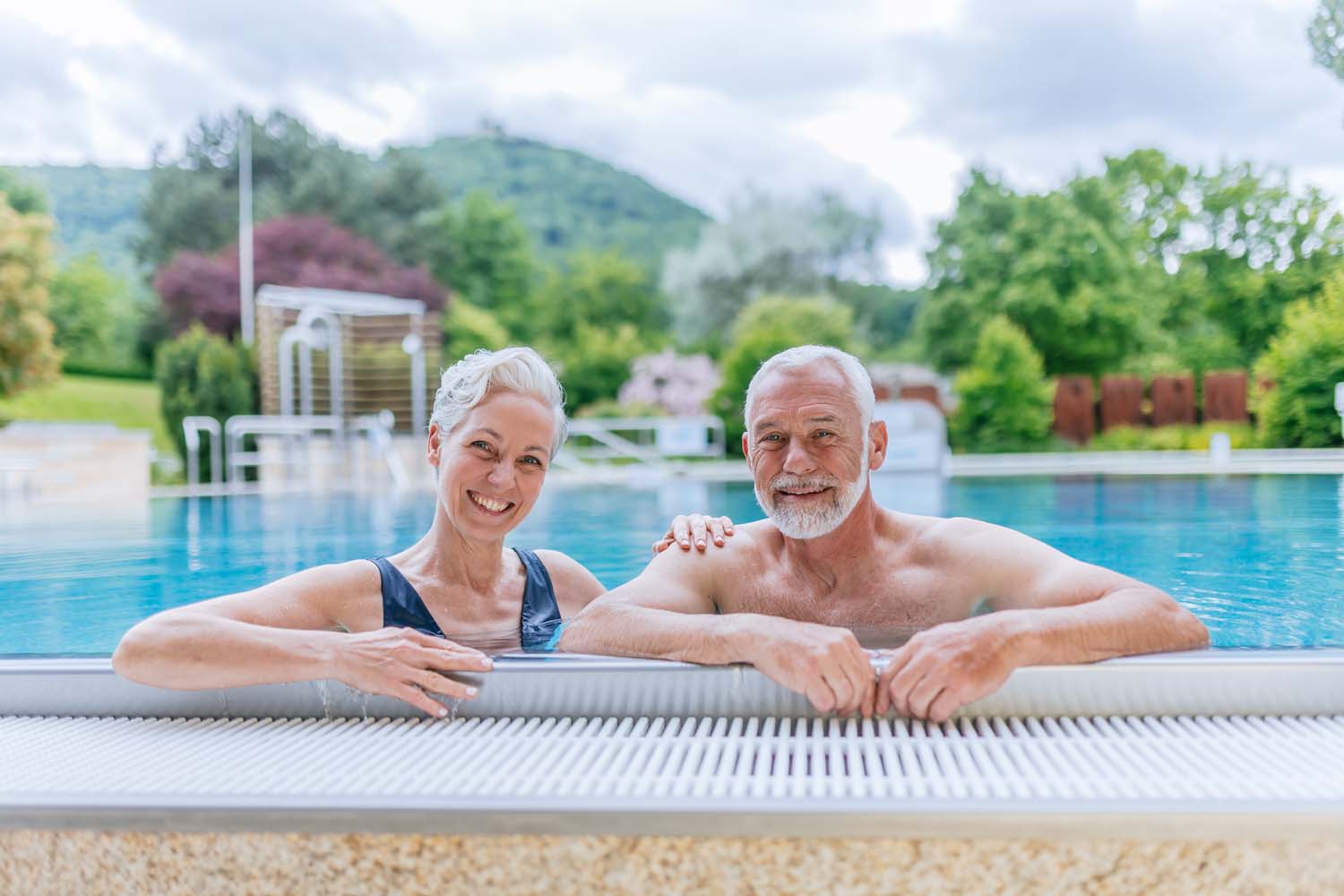 Ein älteres Paar lächelt, während es sich am Rande eines Swimmingpools im Freien entspannt, umgeben von üppigem Grün und Bäumen, mit Hügeln im Hintergrund an einem bewölkten Tag.
