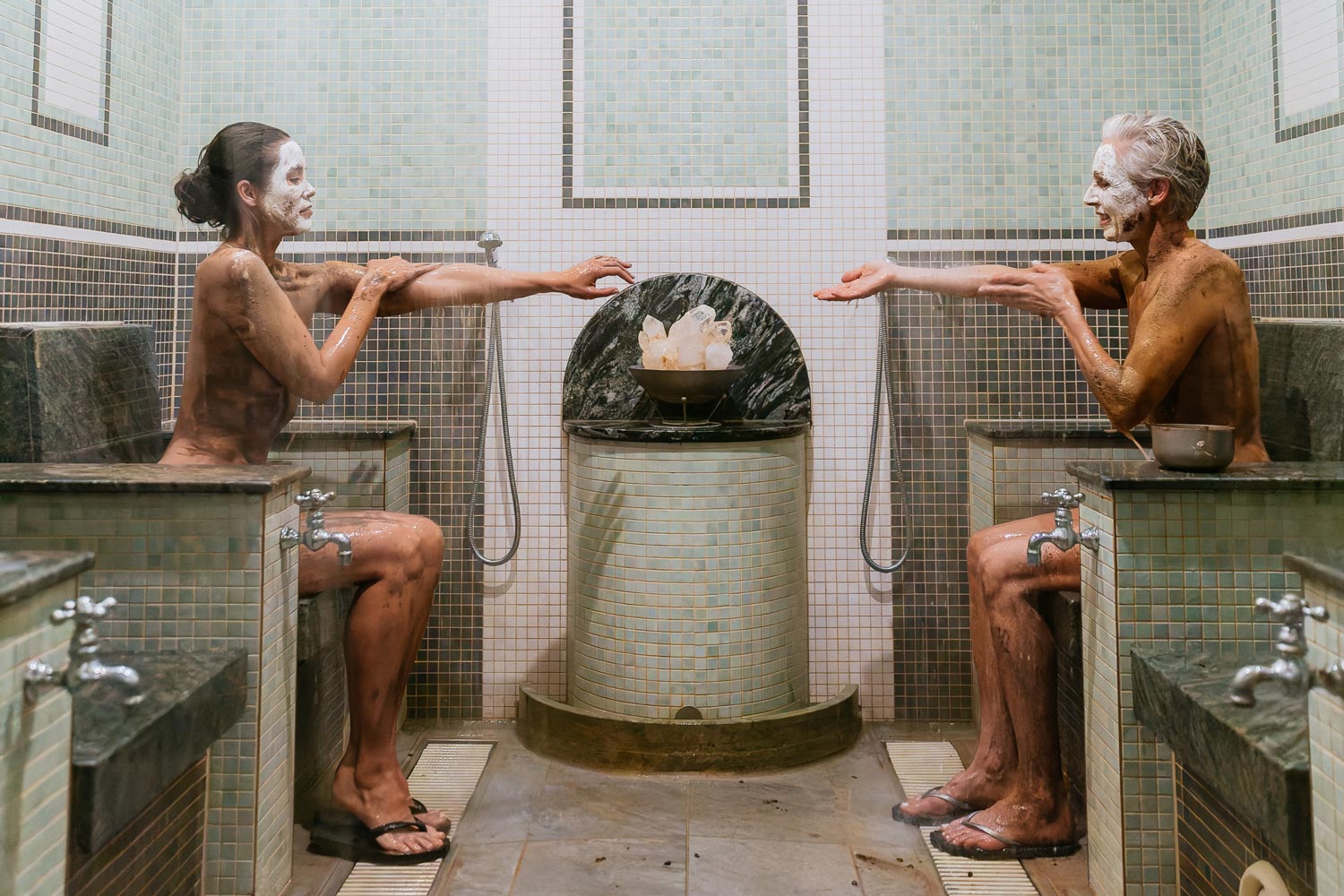 Zwei Personen mit weißen Gesichtsmasken sitzen sich in einem gefliesten Spa-Raum gegenüber und strecken ihre Arme über einen mit Eis gefüllten Marmorbrunnen in der Mitte aus. Sie tragen Sandalen und sind von Spa-Einrichtungen umgeben.