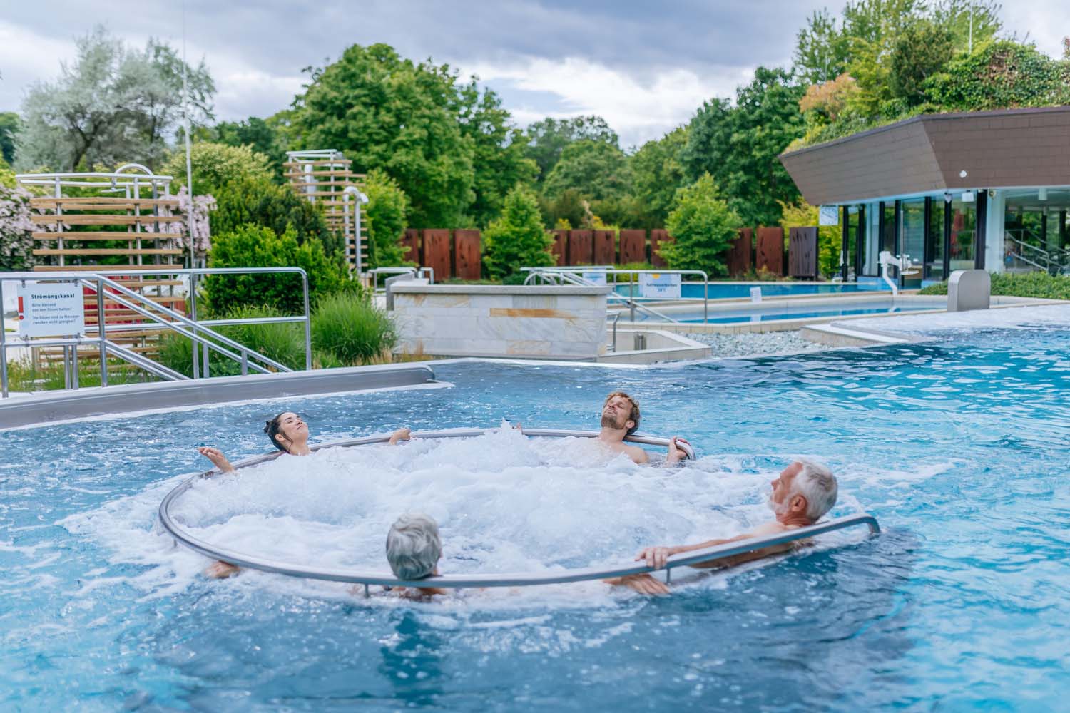 Vier Personen entspannen in einem Whirlpool im Freien mit sprudelndem Wasser, umgeben von Grün und modernen Gebäuden an einem bewölkten Tag.