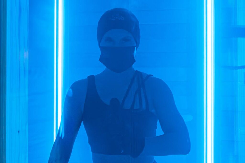Eine Person mit Mütze, Gesichtsmaske und sportlicher Kleidung steht in blauem Licht zwischen zwei vertikalen Lichtröhren, die eine dramatische und futuristische Atmosphäre schaffen.