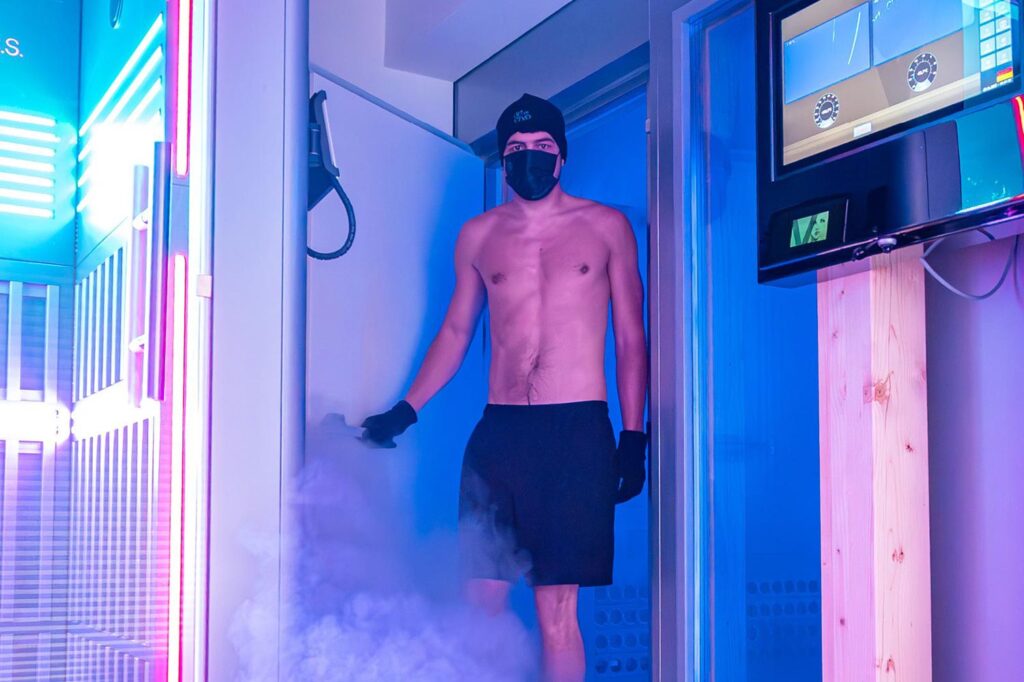 Eine Person ohne Hemd, mit schwarzen Shorts, Handschuhen, einer Mütze und einer Gesichtsmaske steht in einer blau beleuchteten Kryotherapiekammer mit Nebel oder Dampf in der Nähe ihrer Füße. Auf der rechten Seite ist ein Bedienfeld mit Bildschirmen zu sehen.
