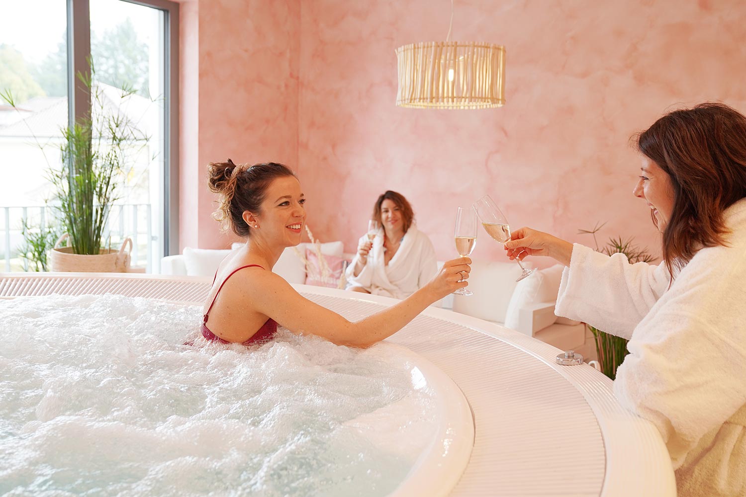 Drei Frauen in weißen Bademänteln entspannen sich in einem Wellness-Raum mit rosafarbenen Wänden; eine sitzt in einem sprudelnden Whirlpool in einem roten Badeanzug und stößt mit einer anderen Frau mit Champagnergläsern an, während die dritte Frau im Hintergrund lächelt.