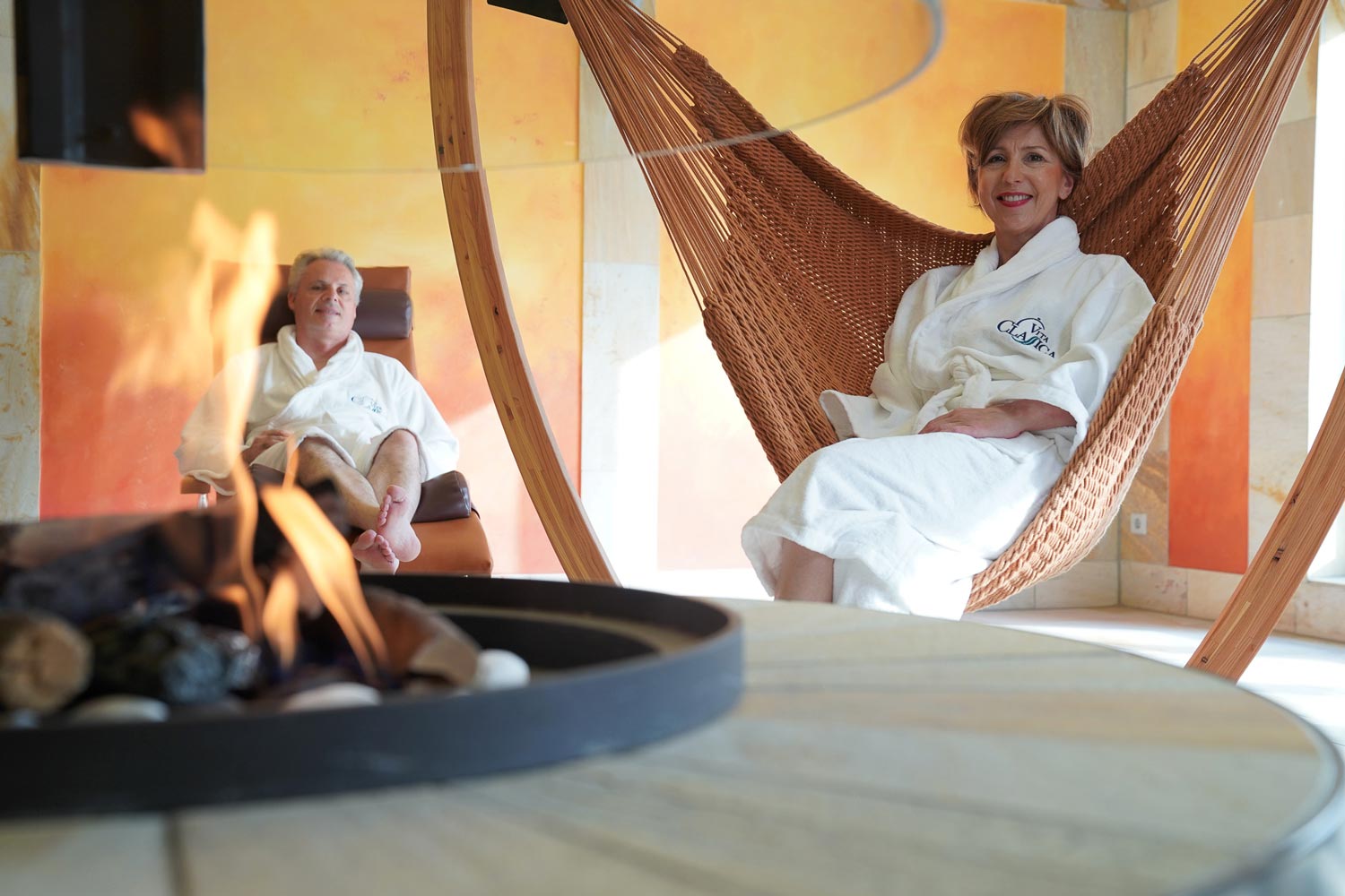 Zwei ältere Erwachsene in weißen Bademänteln entspannen sich an einer modernen Feuerstelle; einer sitzt in einem Hängesessel und lächelt, während der andere im Hintergrund auf einem Liegestuhl Platz nimmt. Der Raum hat warm getönte Wände.