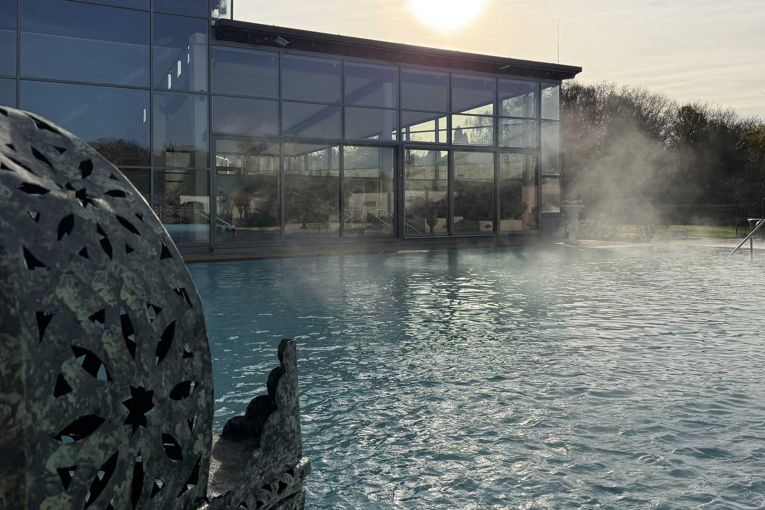 Ein modernes Gebäude mit Glaswänden bietet bei Sonnenaufgang oder Sonnenuntergang einen Blick auf den dampfenden Außenpool der Mediterana Therme, mit Bäumen im Hintergrund und einer verzierten Metalldekoration im Vordergrund.