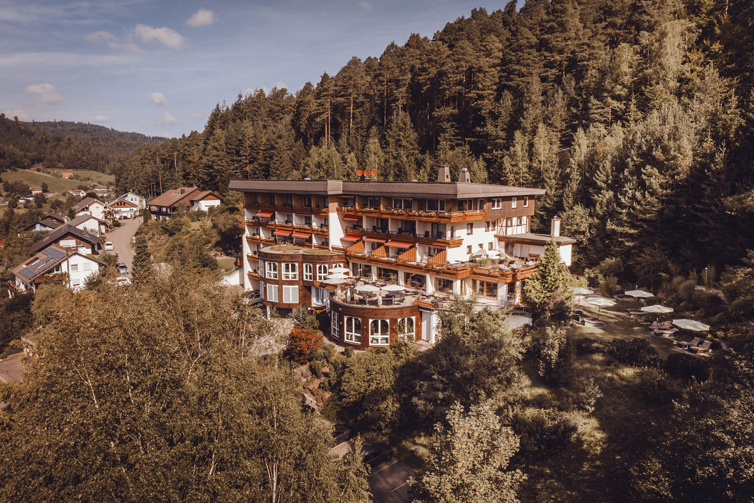 Ein großes, mehrstöckiges Hotel mit Balkonen ist von dichtem Wald und üppigem Grün umgeben und liegt am Rande eines kleinen Dorfes, in dessen Nähe einige Häuser unter einem teilweise bewölkten Himmel zu sehen sind.