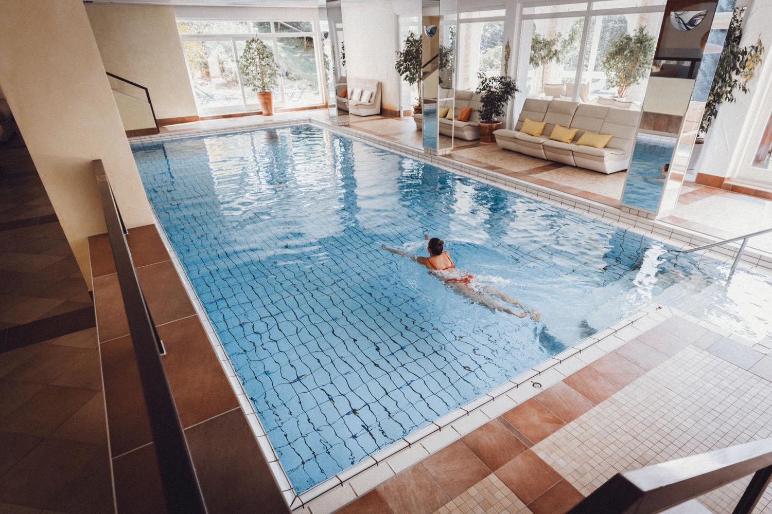 Eine Person schwimmt allein in einem hellen Hallenbad mit großen Fenstern, Topfpflanzen und gepolsterten Sitzen an der Seite. Die Umgebung wirkt ruhig, sauber und entspannend.