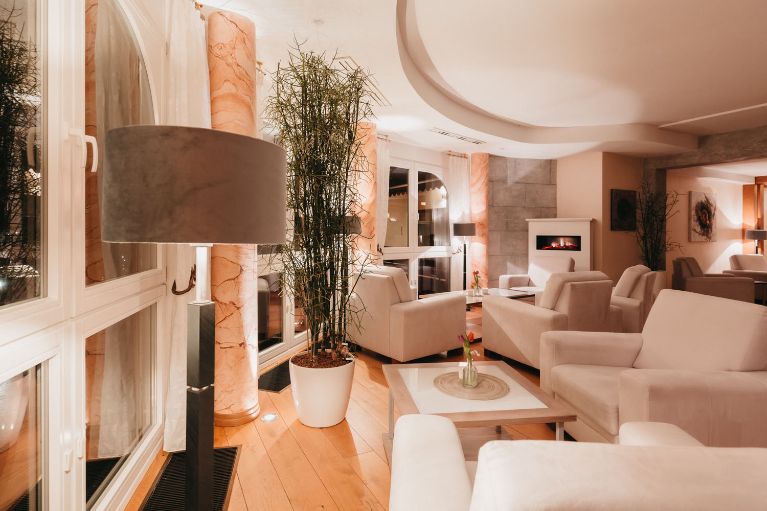 Eine moderne, warm beleuchtete Lounge mit weißen Sesseln, einer grauen Stehlampe, großen Topfpflanzen, Marmorsäulen, Holzfußboden und einem eingebauten Kamin, der eine gemütliche und einladende Atmosphäre schafft.