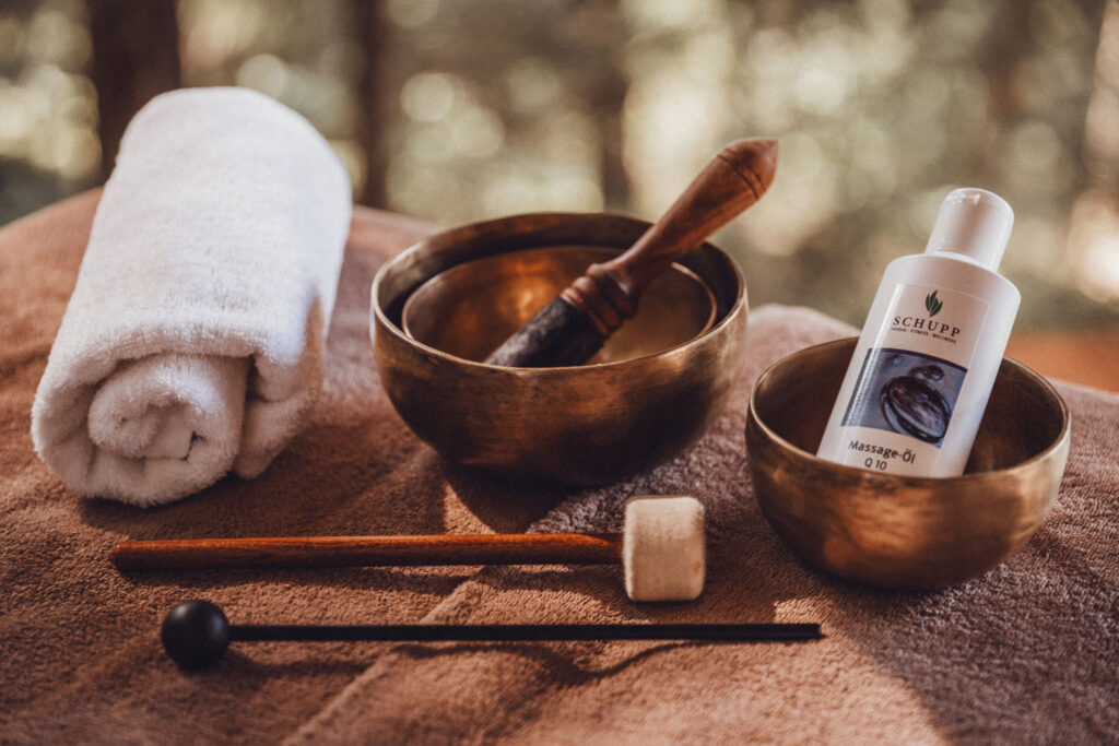 Ein zusammengerolltes weißes Handtuch, zwei Messingschalen - eine mit einem hölzernen Stößel, die andere mit einer Flasche Massageöl - liegen auf einer weichen braunen Oberfläche, davor hölzerne Massagewerkzeuge, vor einem verschwommenen Naturhintergrund.