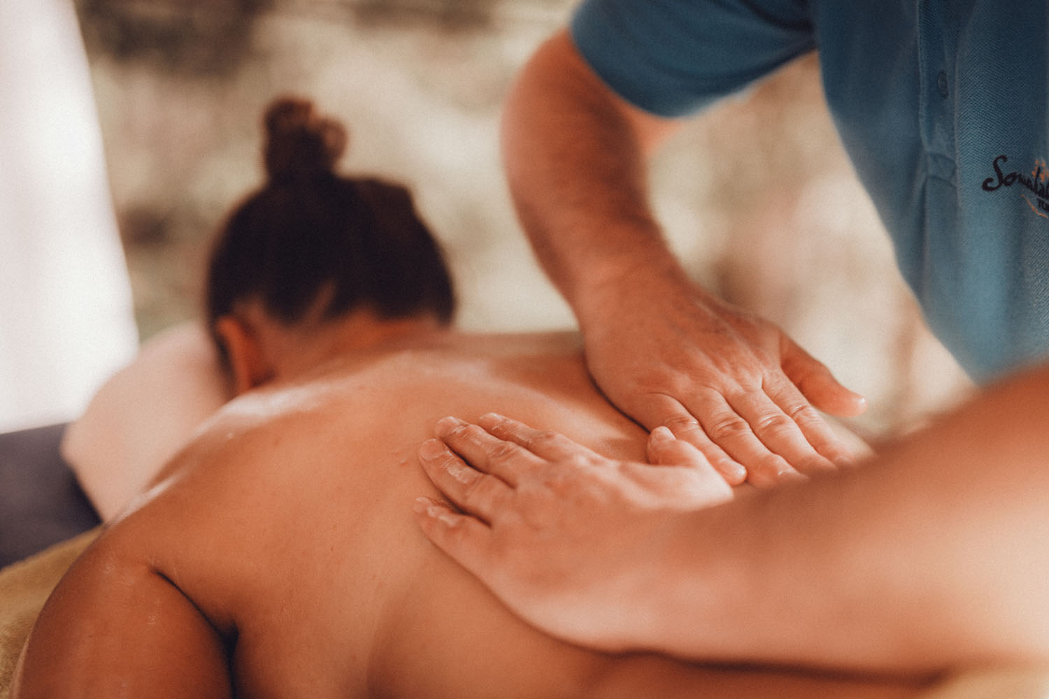 Eine Person erhält eine Rückenmassage von einem Massagetherapeuten, der mit beiden Händen Druck auf den eingeölten Rücken der Person ausübt. Die Person liegt mit dem Gesicht nach unten auf einer Massageliege.