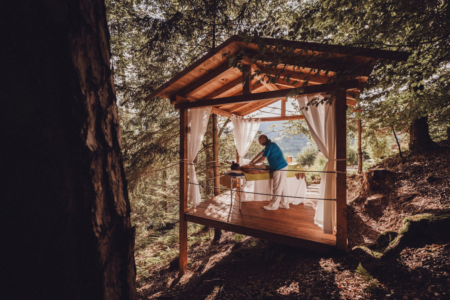 Eine Person erhält eine Massage auf einer hölzernen Plattform mit weißen Vorhängen, umgeben von Bäumen und Natur in einer Waldlandschaft. Ein Massagetherapeut steht neben dem Tisch und arbeitet in der friedlichen Umgebung im Freien.